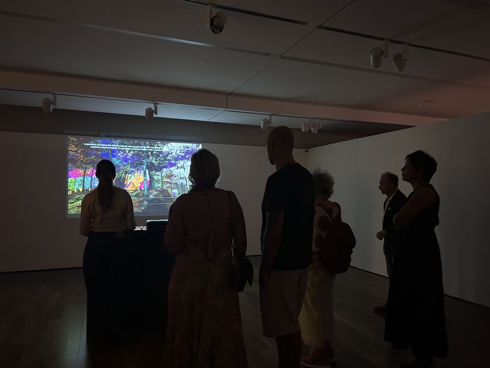 Wide view of the darkened gallery with six visitors gathered in front of the projection