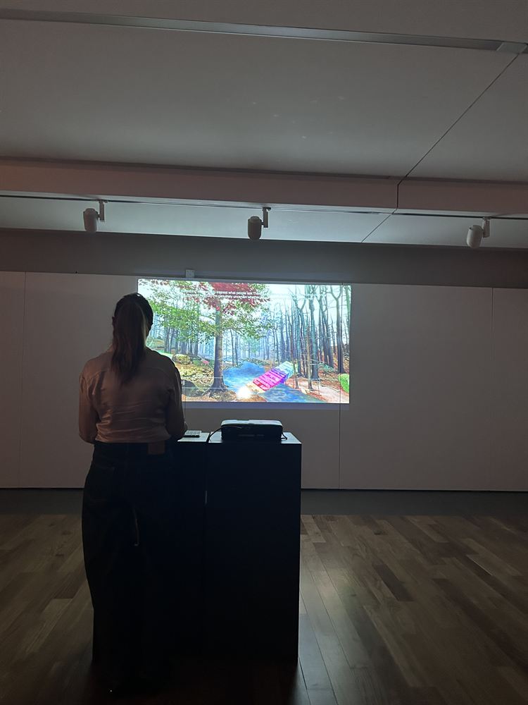 A single visitor alone at the plinth in a wide empty gallery, the forest projected on the far wall