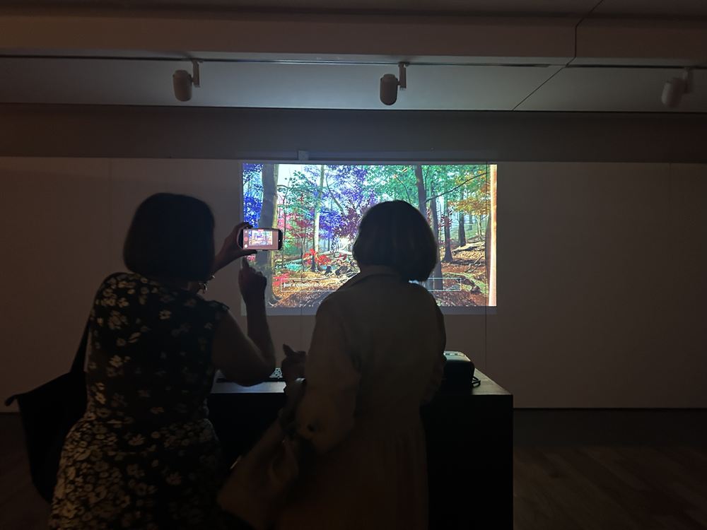 Two visitors at the plinth in a darkened gallery, one typing, the projected forest showing the prompt 'ask a question to AI'