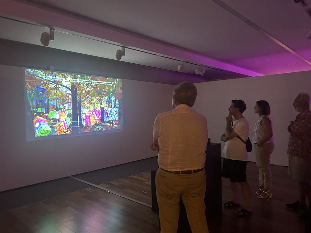 Four visitors watching the projection under magenta gallery lighting, coloured letter blocks piled across the forest floor on screen